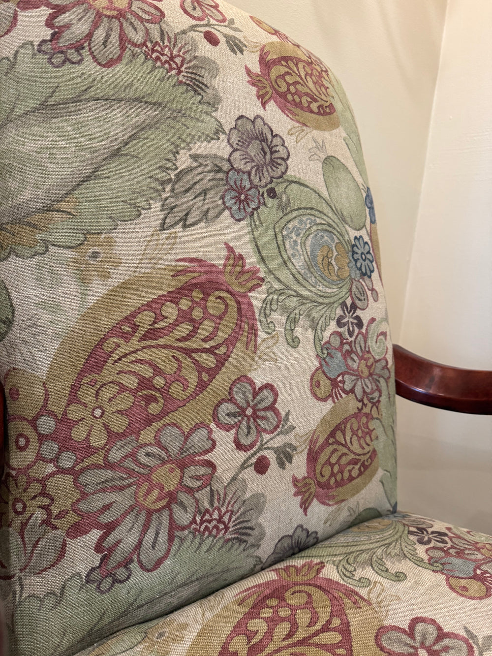 Close-up of upholstered chair fabric with a multicolor floral and paisley pattern in muted green, dusty pink, beige, and blue tones, with a dark wood arm visible on the right.
