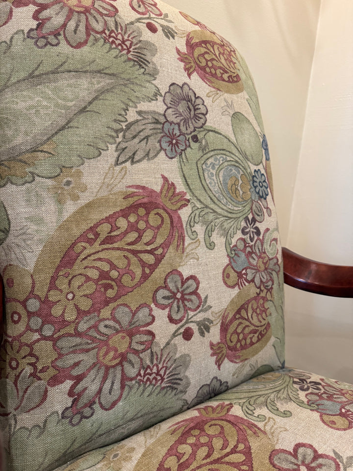 Close-up of upholstered chair fabric with a multicolor floral and paisley pattern in muted green, dusty pink, beige, and blue tones, with a dark wood arm visible on the right.